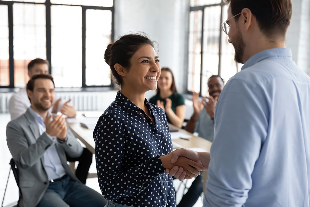 Two colleagues shaking hands while others clap in the background