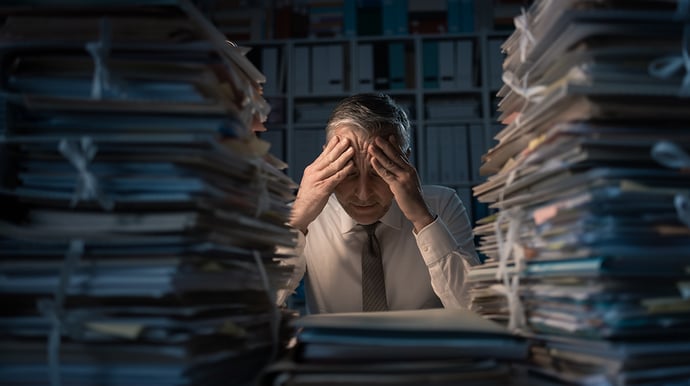 Overworked Employee with his hands on his head, surrounded by mountains of paperwork on his desk.